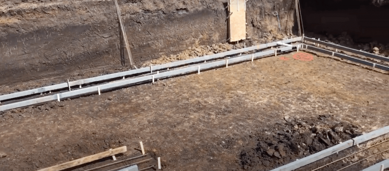 Excavation site with a large, empty dirt area surrounded by retaining walls. Metal beams define the rectangular perimeter, and construction materials are scattered in the foreground. The background shows a plywood-covered section and a green tool hanging on the wall.