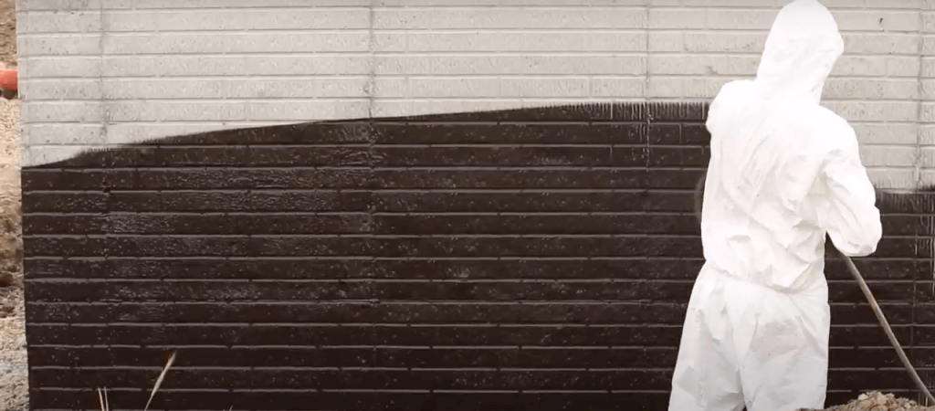 A person in a white protective suit and hood paints a brick wall with black paint using a long-handled roller. The upper part of the wall remains white, while the lower part is freshly coated black.