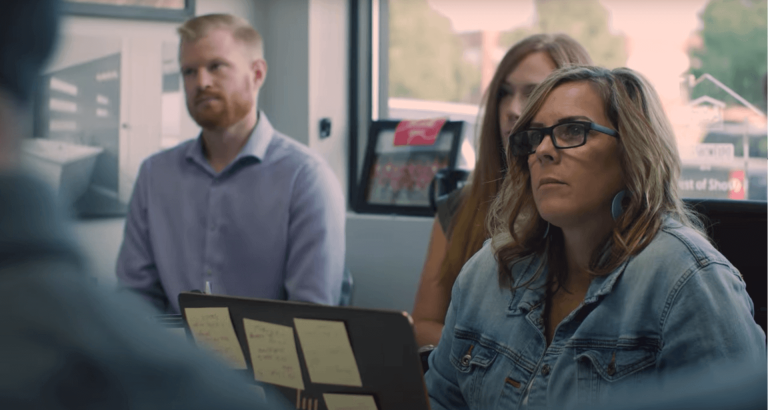 A group of three individuals, consisting of two women and one man, sits attentively during a meeting in a well-lit office. The woman in the foreground wears glasses and a denim jacket, with a laptop in front of her displaying sticky notes.