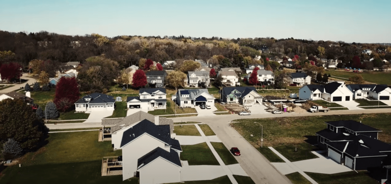 Aerial view of a suburban neighborhood featuring several houses with yards, trees with autumn foliage, and empty plots where houses are under construction. An empty road runs through the area, and the background shows a mix of forested and residential areas.