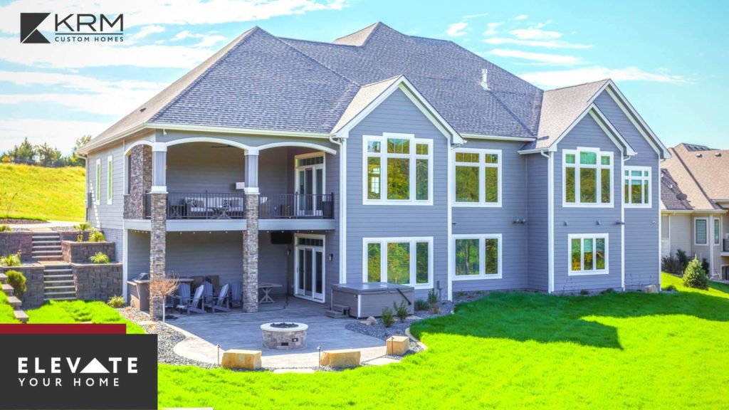 A large, modern, gray two-story home with multiple windows, a spacious balcony, and a covered patio area is shown. The backyard has green grass and a round stone fire pit. The logo "KRM Custom Homes" and the tagline "Elevate Your Home" are displayed on the image.