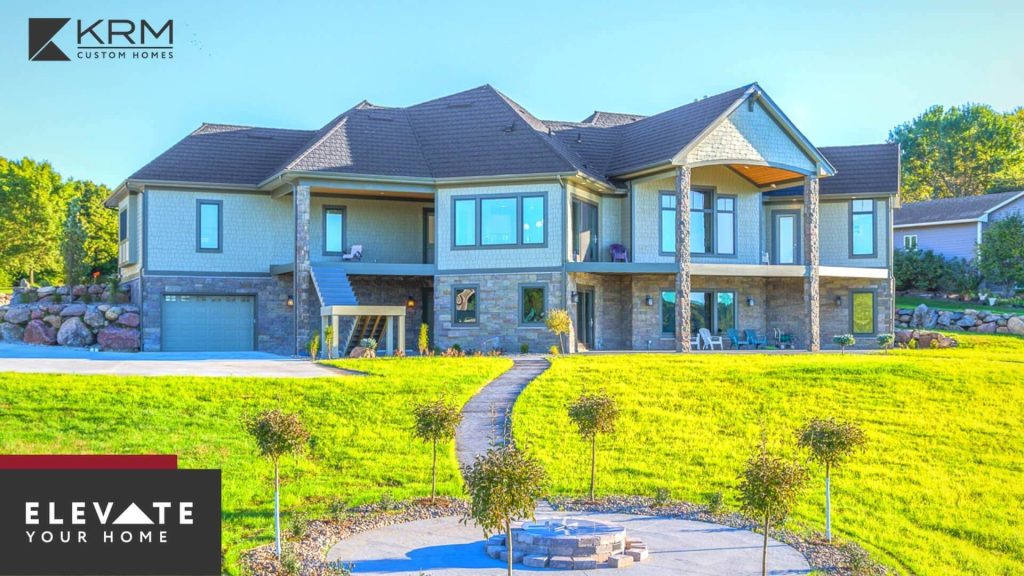 A large, two-story custom home with multiple gables and a mix of stone and siding exteriors. A curved pathway leads to the entrance, surrounded by a well-manicured lawn and young trees. The logo "KRM Custom Homes" and the tagline "Elevate Your Home" appear in the corners.