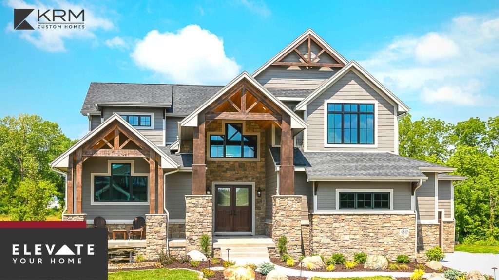 A modern, two-story house with grey siding, large windows, and wooden beams framing the entrance. The home features a neatly landscaped front yard with shrubs and stone accents. The logo for KRM Custom Homes and the text "Elevate Your Home" are positioned in the lower left corner.