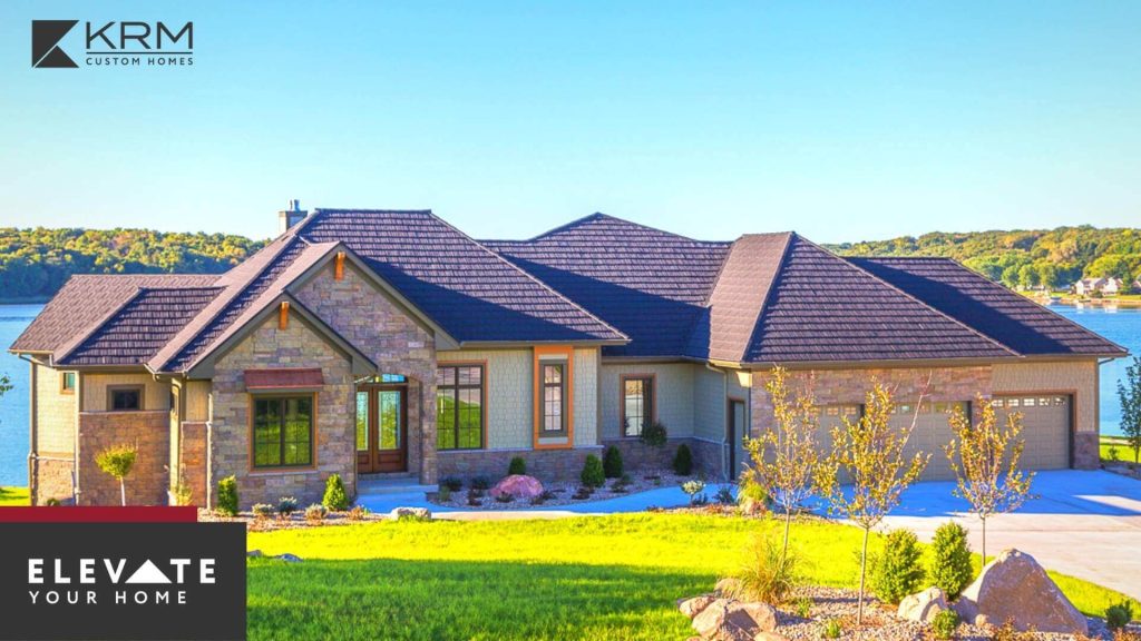 A modern, single-story house with a stone and wood exterior sits on a lush green lawn overlooking a large body of water. The home features a dark, shingled roof and multiple large windows. The text "KRM Custom Homes" and "ELEVATE YOUR HOME" appear in the corners.