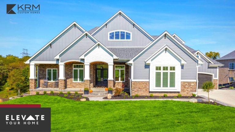 A modern two-story house with a gray and white exterior, stone accents, and a well-maintained green lawn. The house features multiple gables, large windows, and an inviting front porch. The logo "KRM Custom Homes" is in the upper left, and "ELEVATE YOUR HOME" is in the lower left.