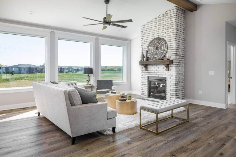 Modern living room with large windows, gray sofa, white brick fireplace, and wooden floor.