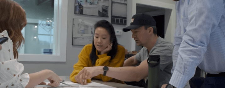A group of four people are gathered around a table in a modern office space, discussing a document. One person in a grey shirt and black cap is pointing at the paper while another in a mustard yellow sweater is attentively leaning in. The background shows framed pictures on the wall.