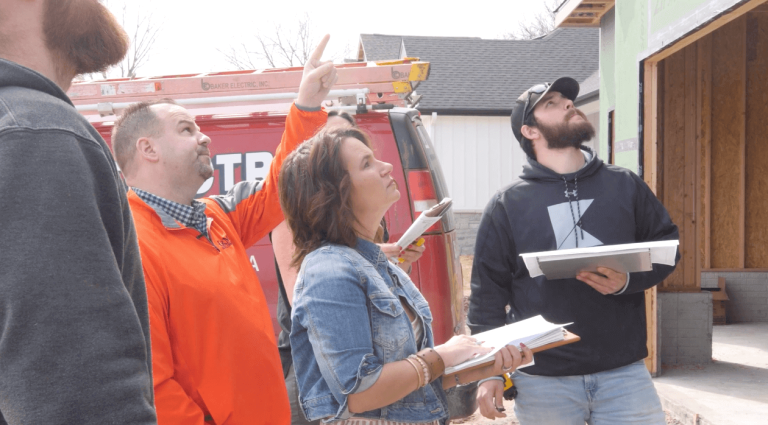 A group of four people standing outside a partially constructed building. One person in an orange shirt points upward while holding papers. Another person in a denim jacket looks up attentively, and two others hold folders, observing the structure.