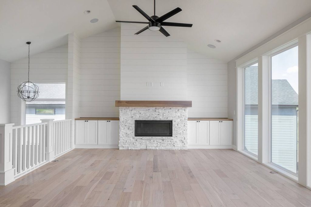 A modern, white living room with a vaulted ceiling, featuring a central fireplace with a stone surround. The fireplace is flanked by built-in cabinets. A large window fills the right wall, and a ceiling fan with dark blades hangs in the center of the room.