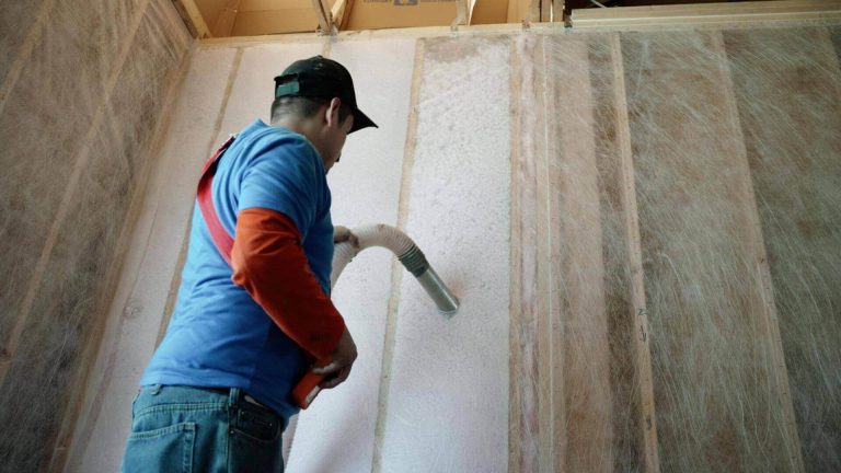 A worker, wearing a cap and a blue shirt with an orange sleeve, holds a hose and blows insulation into the gaps between wall studs during a home construction or renovation project. The wall frame is visible, and the insulation appears white and fluffy.