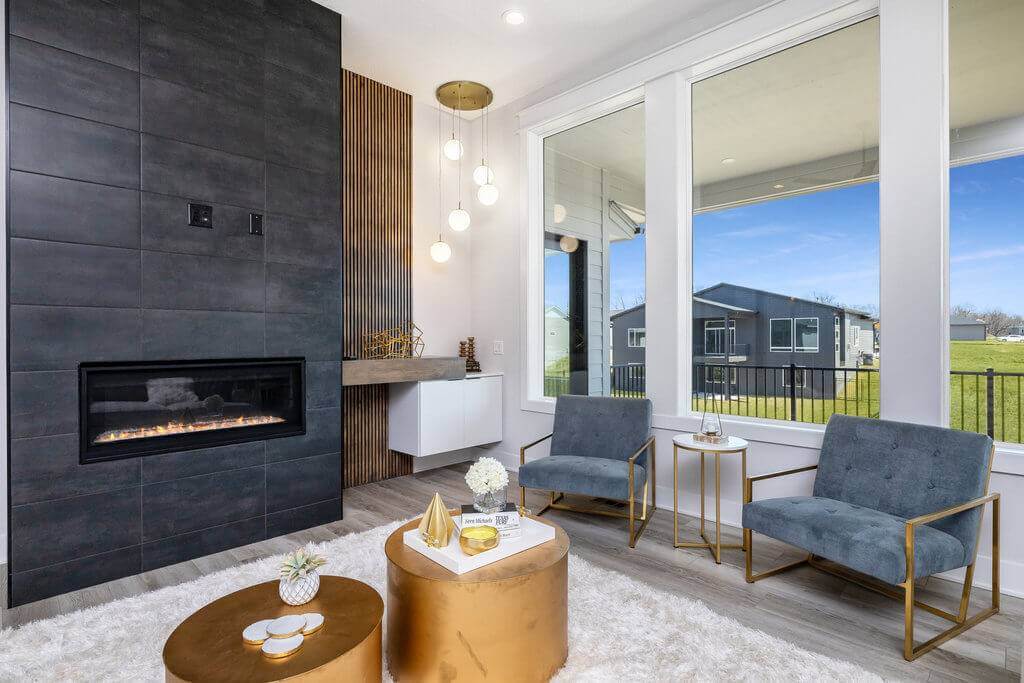 A modern living room featuring a sleek black tiled fireplace with a cozy fire. Two gray armchairs with gold frames are positioned around a round, gold coffee table. A large window offers a clear view of a green lawn and house outside. Hanging pendant lights add warmth.