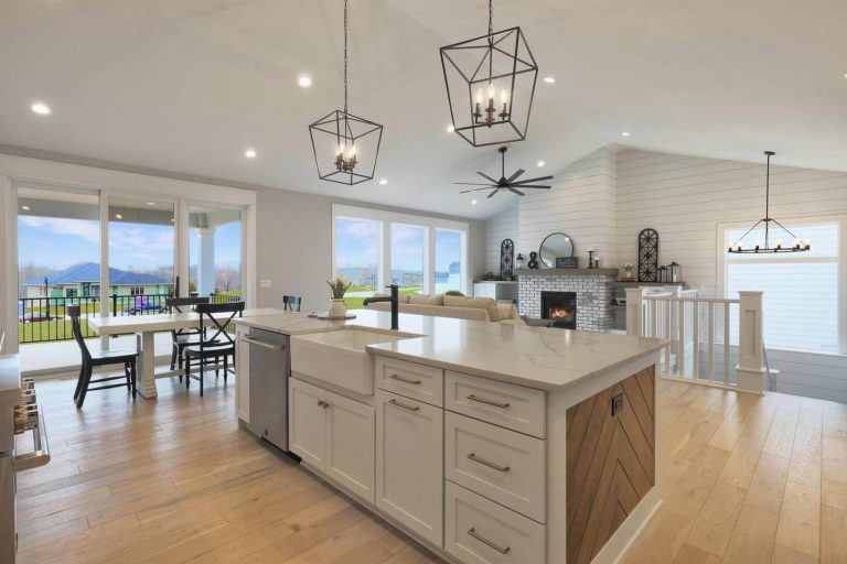 A modern open-concept kitchen and living area with light wood floors. The kitchen features a large island with a farmhouse sink and pendant lights. The living room has a fireplace and shiplap walls. Large windows and a dining area offer scenic views of mountains and greenery.