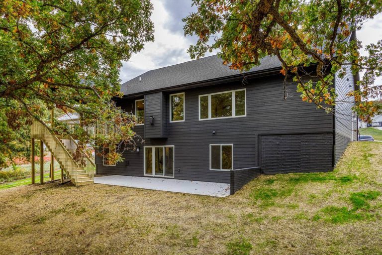 Modern dark gray house with large windows, back patio, and stairs, surrounded by trees and grass.