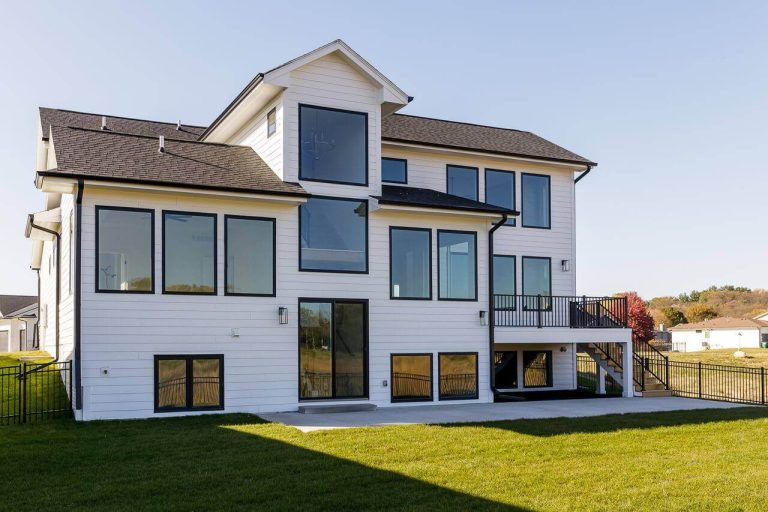 A modern, multi-story house with white siding and large windows. The house features a dark roof and a spacious backyard with a manicured lawn. A staircase leads from an elevated deck to the lawn. The sky is clear, and neighboring houses are visible in the distance.