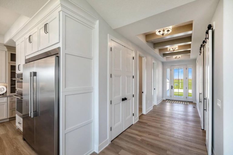 A modern hallway with light wood flooring and white walls, featuring recessed ceiling lights and exposed beams. A large stainless steel refrigerator is visible on the left, adjacent to a kitchen area. The hallway leads to a glass door with a view of greenery outside.
