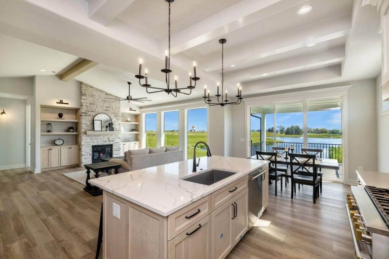 Modern open-concept kitchen and living room with a large marble island, farmhouse sink, and elegant lighting fixtures. The living area features a stone fireplace and built-in shelving. Large windows and sliding doors offer a scenic view of a lake and greenery.