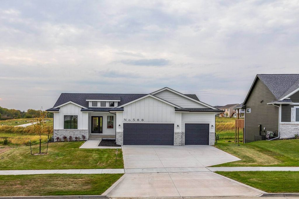 A modern single-story house with light gray siding and white accents. The front features a triple garage, large windows, and a well-maintained lawn. The house number "588" is visible above the garage. Sky is overcast, with other houses and grassy fields in the background.