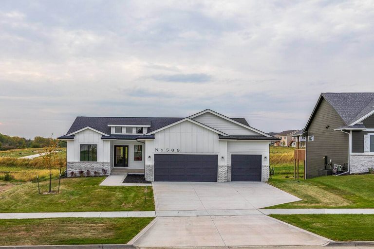 A modern, single-story house with a white exterior, gray roof, and three-car garage. The home features a neatly manicured lawn and is set against a cloudy sky backdrop. The house number 588 is displayed above the garage. Houses and greenery are visible in the background.