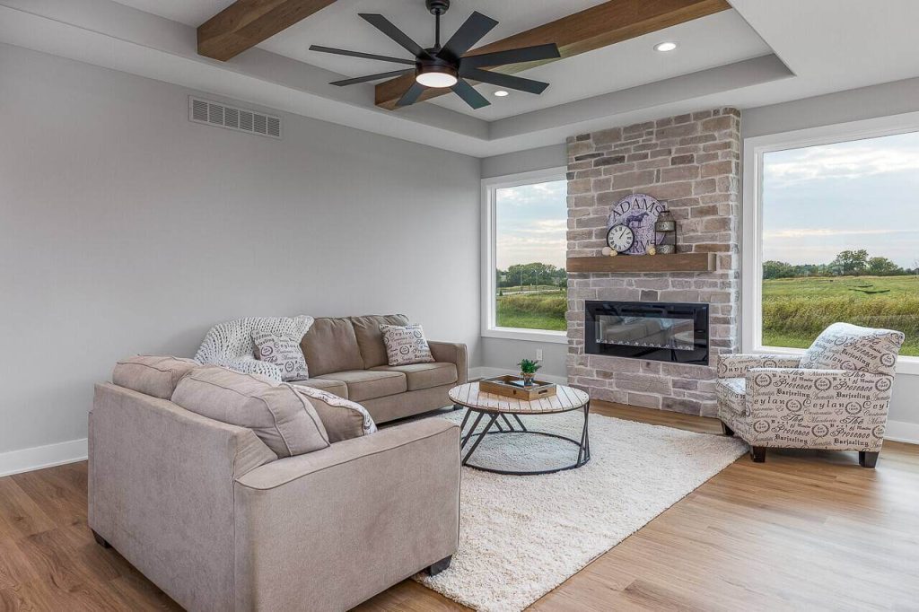 A modern living room with a large window showing an outdoor view. The room features two beige sofas, a patterned armchair, a round wooden coffee table, and a brick fireplace with a clock on the mantel. A ceiling fan with wooden beams is overhead, and a rug covers the wooden floor.