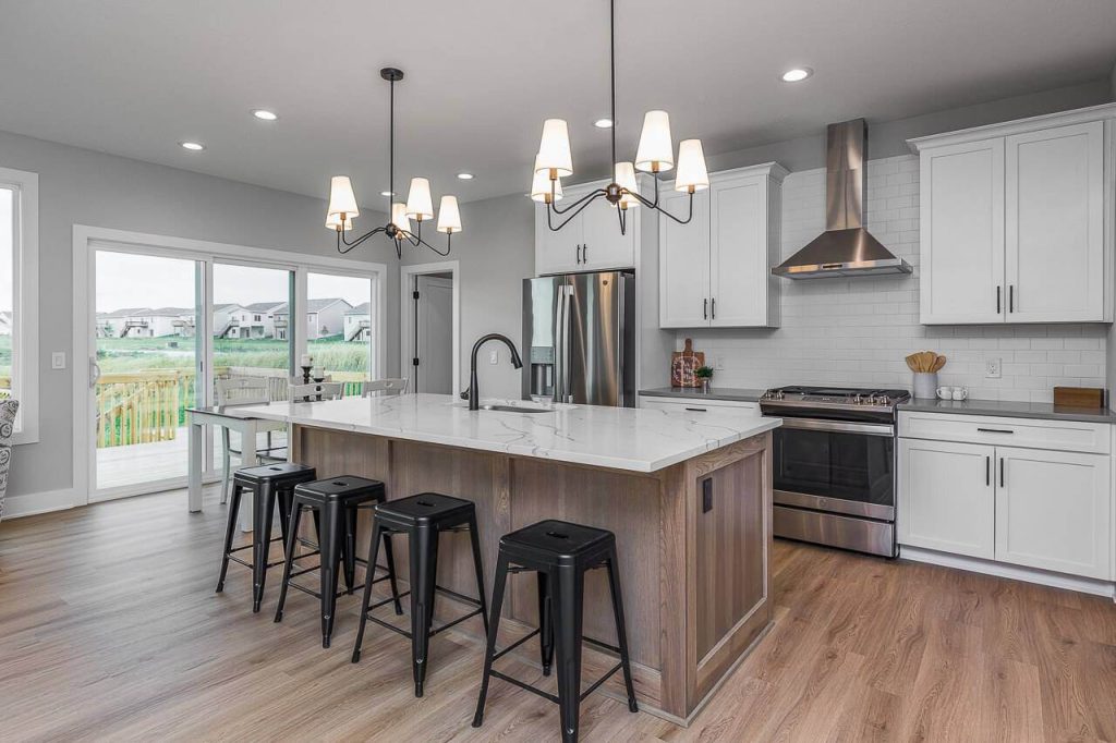 Modern kitchen with a large central island featuring a white marble countertop and four black bar stools. White cabinets, stainless steel appliances, and two pendant lights above the island enhance the sleek look. Glass doors open to a deck with a view of the backyard.