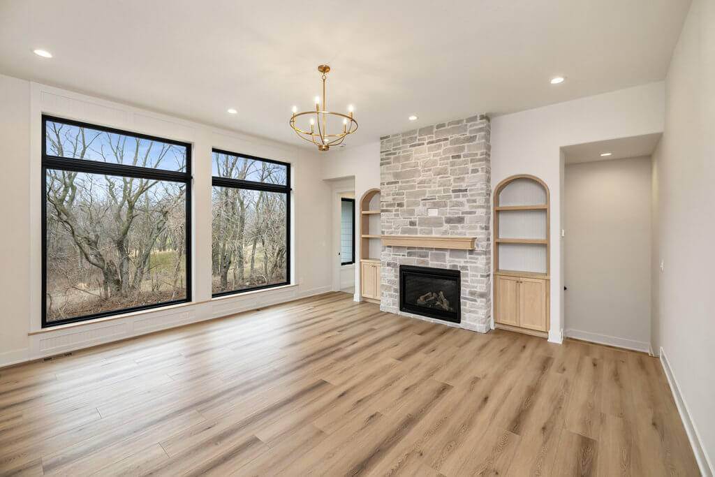 A spacious living room with light wood flooring, a stone fireplace, built-in shelves on both sides, three large windows with black frames providing a view of trees, and a modern chandelier hanging from the ceiling. The room is well-lit with recessed lighting.