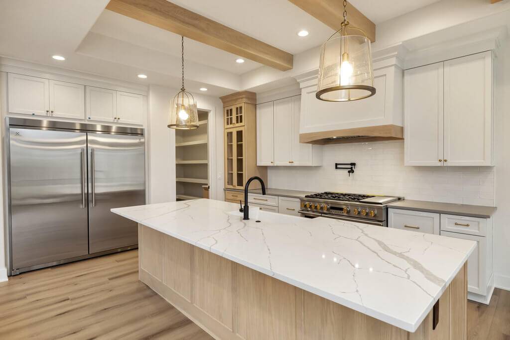 A modern kitchen featuring a large central island with a white marble countertop, a black faucet, and pendant lighting overhead. The kitchen includes stainless steel appliances, white cabinetry, and light wooden accents, including beams on the ceiling.