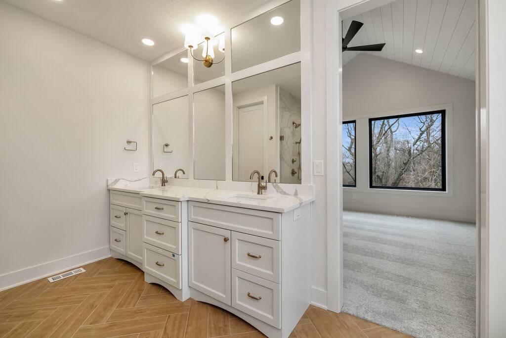 A modern bathroom with double vanity sinks featuring marble countertops and white cabinetry beneath large mirrors. Golden fixtures and handles adorn the sinks. To the right is an open doorway leading to a bedroom with large windows and a view of trees outside.