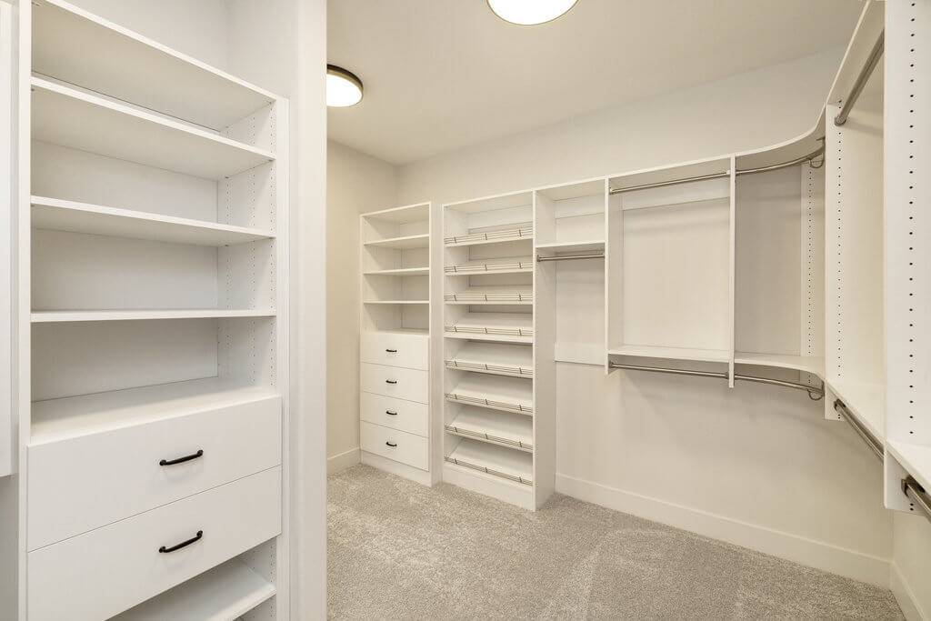 Spacious walk-in closet with white built-in shelving, drawers, and hanging rods. The room is well-lit with ceiling lights and features a light beige carpet. Various shelves and drawers provide ample storage space.