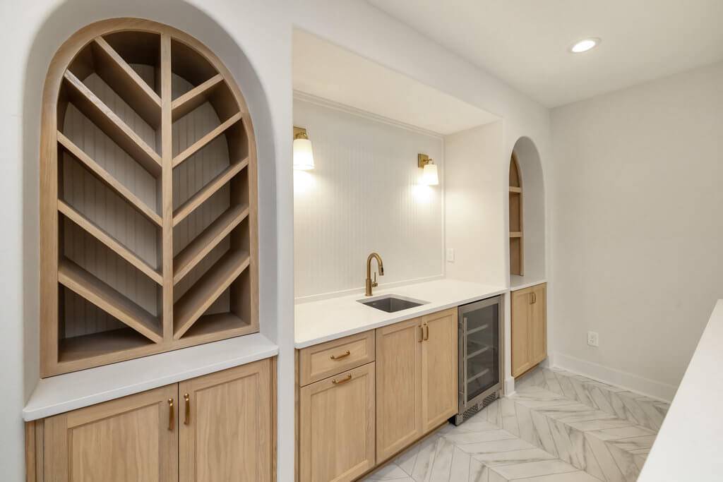 A modern pantry with light wooden cabinetry and white countertops. It features a chevron-patterned wine rack, a sink with a brass faucet, and a small built-in fridge. The space is illuminated by two wall-mounted lights. The floor has a herringbone tile pattern.