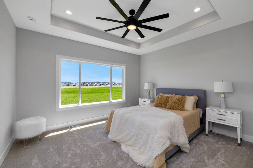 A modern bedroom with a large window overlooking a grassy exterior. The room features a bed with a beige comforter and fluffy pillows, flanked by two white nightstands with gold-accented lamps. A white furry stool sits in the corner, and a ceiling fan hangs above.