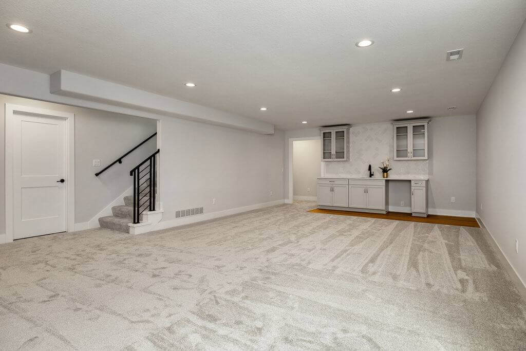 A spacious, carpeted basement features a staircase with a black handrail, recessed lighting, and light gray walls. On one side, there is a small kitchenette with white cabinets, a countertop, and a few decorative items.
