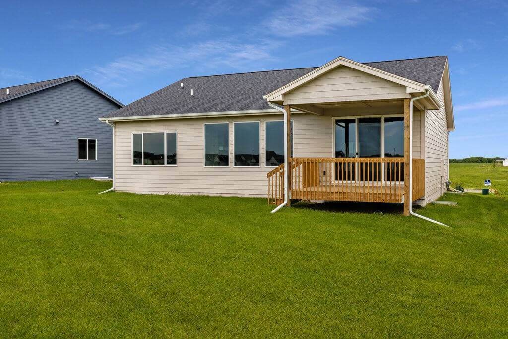 A single-story house with light-colored siding and a dark shingled roof. The house features large windows and a small covered wooden porch at the back. It sits on a spacious, well-maintained grass yard under a clear blue sky.