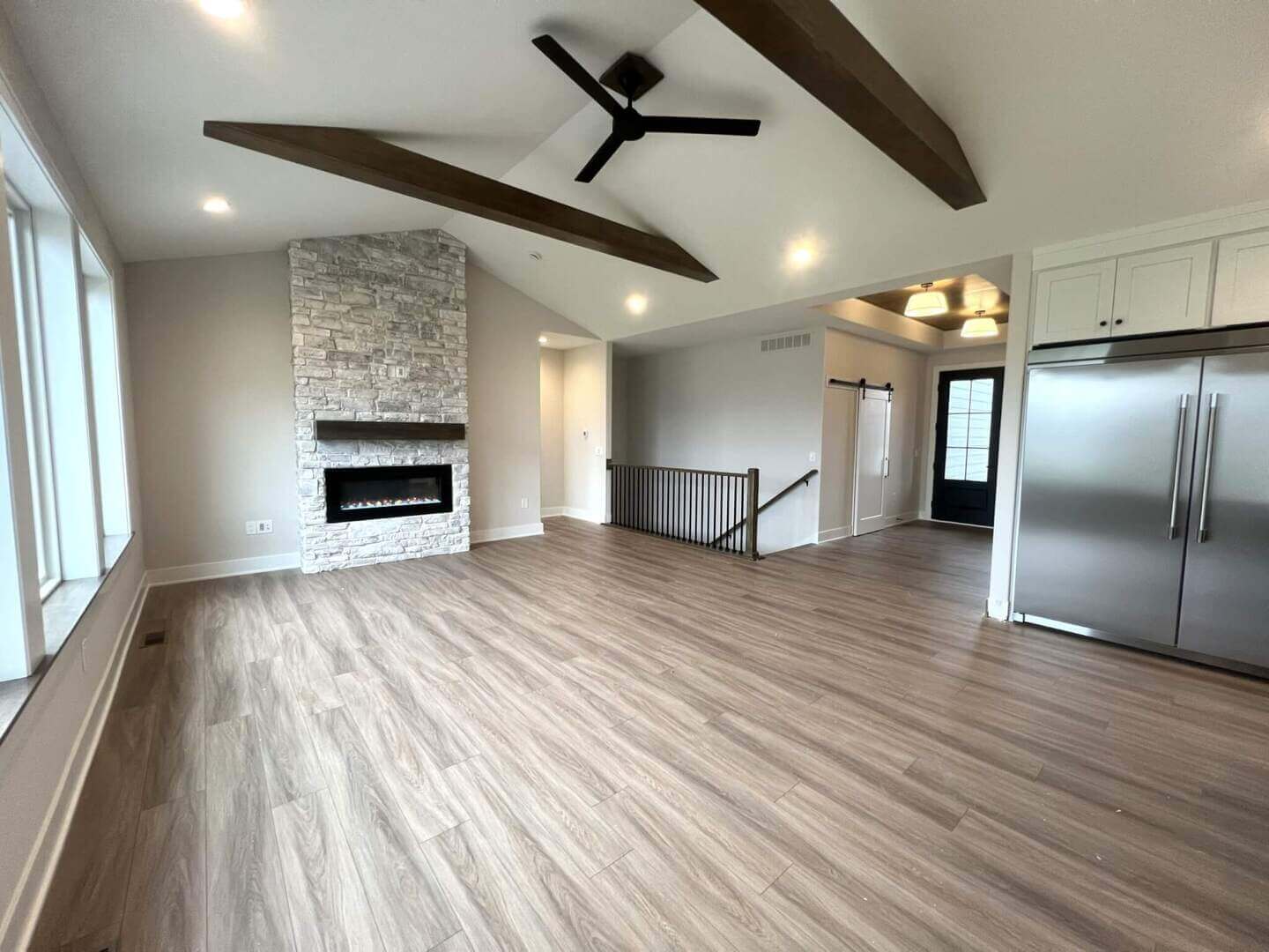 A spacious, modern living area with light wood flooring and a tall ceiling featuring wooden beams and a black ceiling fan. The room includes a stone fireplace, a large window, and a stainless steel refrigerator next to white cabinets. A staircase leads downstairs.