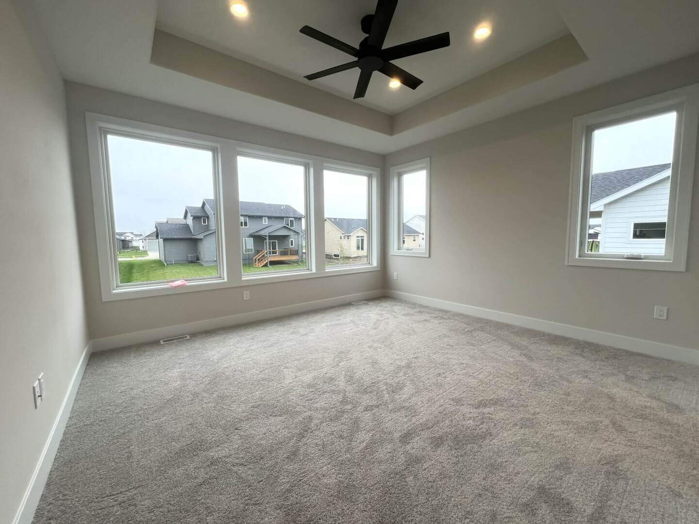 A spacious, empty room with beige walls and carpeted flooring. The room has large windows on two sides, allowing natural light to flood in. The ceiling features recessed lighting and a modern black ceiling fan. Outside, a suburban neighborhood is visible.
