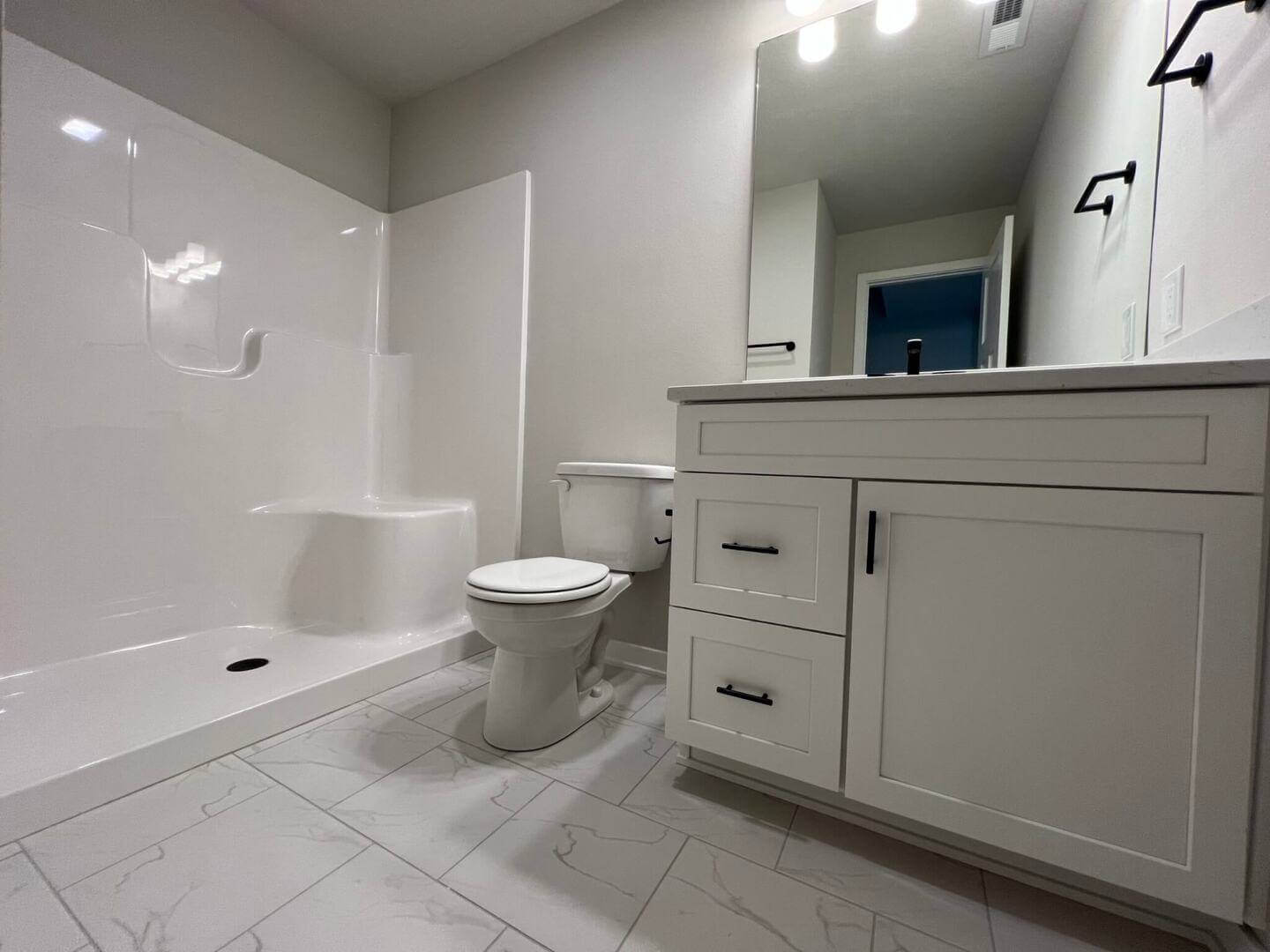 A modern bathroom featuring white fixtures, including a shower with built-in shelves, a toilet, and a vanity with a large mirror and two-light fixture above. The floor is tiled in a light color, and the vanity has black hardware on the drawers and cabinets.